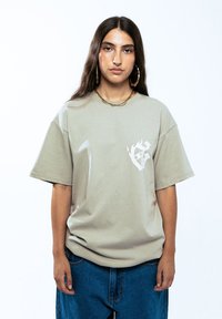 Jasnoszary, oversize t-shirt z krótkim rękawem, z małym białym wzorem graficznym na lewej stronie klatki piersiowej. Materiał bawełniany.