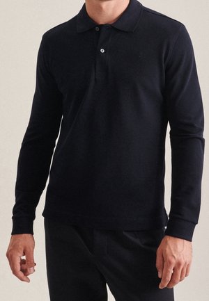 Mann trägt ein schwarzes Poloshirt mit langen Ärmeln und zwei Knöpfen, steht mit entspannten Armen an den Seiten.