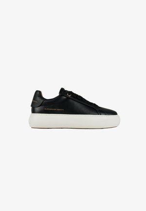 Zwarte lage leren sneaker met dikke witte zool, zwarte veters en gouden "Alexander Smith"-branding aan zijkant en hiel.