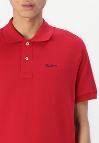 Rdeča polo majica iz teksturiranega materiala, z ostrim ovratnikom, dvema gumboma in modrim logotipom vezenim na prsih.