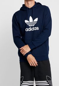 Person som bär en marinblå Adidas-hoodie och svarta Adidas-shorts, står med armarna i kors mot en enkel ljus bakgrund.
