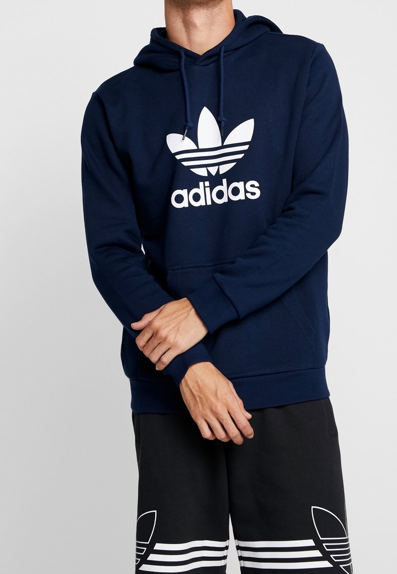 Person som bär en marinblå Adidas-hoodie och svarta Adidas-shorts, står med armarna i kors mot en enkel ljus bakgrund.