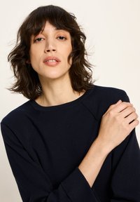 Frau mit schulterlangem, welligem braunem Haar, die ein dunkelblaues Langarmoberteil trägt und mit der linken Hand ihre rechte Schulter hält.