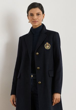Donna che indossa un cappotto blu navy scuro con bottoni dorati e uno stemma ricamato sulla tasca del petto, abbinato a un dolcevita nero e orecchini.