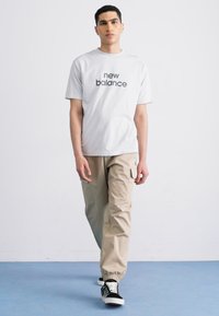 Weißes T-Shirt mit "New Balance"-Logo, kombiniert mit beigen Cargo-Hosen und schwarz-weiß gemusterten Sneakers. Einfaches, lässiges Ensemble.