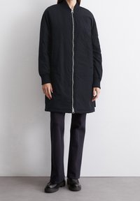 Osoba ubrana w długą czarną kurtkę na zamek, ciemne spodnie i czarne buty, stojąca na tle jednolitej jasnej ściany i podłogi.