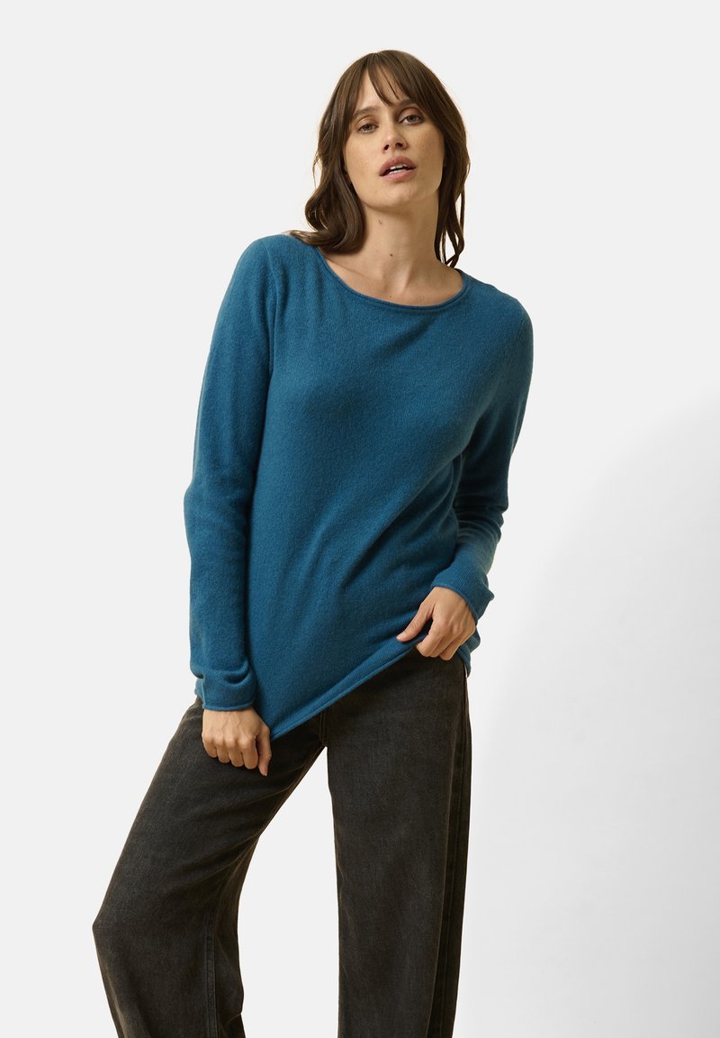 Suéter turquesa de manga larga con un escote ancho y puños acanalados, combinado con jeans oscuros de pierna ancha. Textura suave y corte relajado.