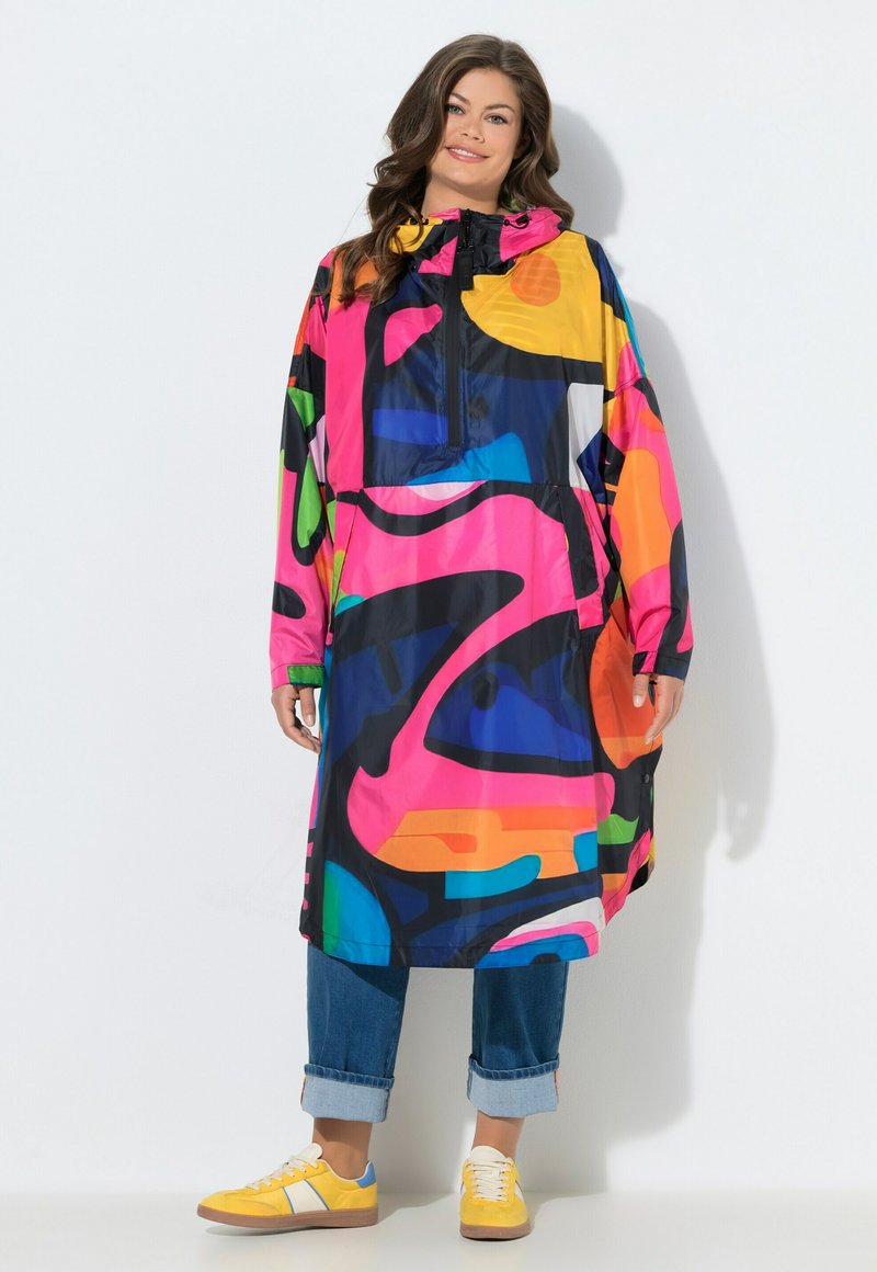 Vrouw draagt een kleurrijke lange jas met abstract patroon en capuchon, blauwe opgerolde spijkerbroek en gele sneakers, staand tegen een witte achtergrond.