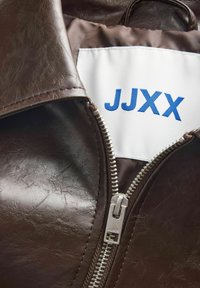 Hnědá faux kůže bunda s šedým zipem a bílou etiketou s modrým textem "JJXX." Texturovaný povrch s mírným leskem.