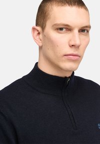 Navy zip-up sweater with high collar, knit texture, and a small logo on the chest. Close-fitting design.
