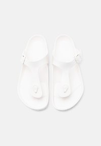 Ζευγάρι λευκών σανδαλιών slip-on με διαχωριστικό δαχτύλου και ρυθμιζόμενα λουριά με αγκράφα σε απλό ανοιχτό φόντο.