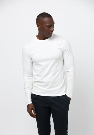 Langærmet T-shirt med rund hals i blødt hvidt stof med en glat tekstur, parret med mørke bukser, stående mod en ensfarvet baggrund.