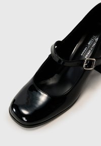 Black patent leather Mary Jane shoe with a square toe, an adjustable strap, and a small silver buckle accent. Vegan-friendly.