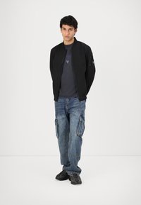 Black bomber jacket, gray t-shirt with logo, blue cargo jeans, and black sneakers. The jacket features a zip closure and shoulder pocket.