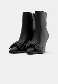 Bottines noires à bout pointu, talons hauts, fermeture éclair latérale et grands nœuds décoratifs avec détails dorés à l'avant.
