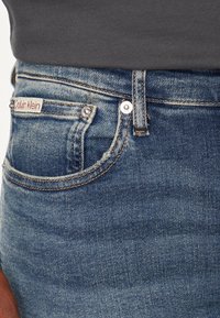 Vaqueros de denim de Calvin Klein con un acabado azul desgastado, que cuentan con un bolsillo frontal, herrajes plateados y costuras en contraste en color beige.