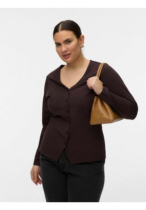 Sötétbarna gombos blúzt és fekete nadrágot viselő nő, vállán bézs bőrtáskát hord, egyszínű világos háttér előtt.