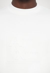 Camiseta de algodón blanca con un logotipo texturizado en relieve que muestra "ARMANI" y "EXCHANGE" en gris, cuello redondo y mangas cortas.