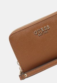 Cartera de textura marrón con herrajes dorados y logotipo en relieve "GUESS EST 1981". Cierre con cremallera en el borde superior, de forma rectangular delgada.