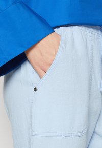 Camisa azul claro con tejido texturizado, que presenta un bolsillo con un botón de presión negro. La manga cuenta con un puño de tela azul de contraste.