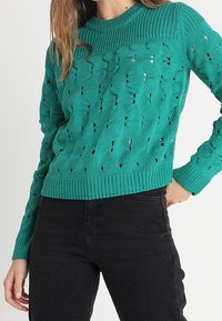 Turkos stickad tröja med ett texturerat mönster och hålmönsterdetaljer, med en ribbad rundhals och åtsittande ärmar, som är kombinerad med svarta byxor.