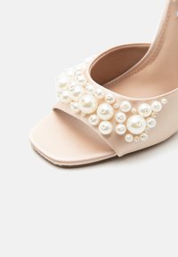 Μπεζ γόβα με ανοιχτό δάχτυλο και λουράκι διακοσμημένο με λευκές πέρλες διαφόρων μεγεθών και μικρές ασημένιες λεπτομέρειες.