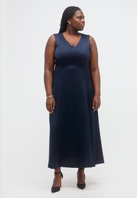 Γυναίκα με μακριά κοτσίδα που φοράει αμάνικο μακρύ μπλε φόρεμα και μαύρα τακούνια, στέκεται και κοιτάζει προς το πλάι μπροστά σε λευκό φόντο.