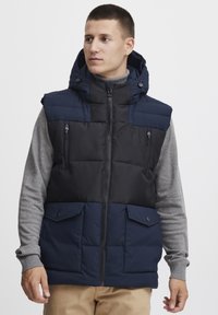Navy and black puffer vest with a hood, zippered pockets, and buttoned front, worn over a gray long-sleeve shirt. Textured fabric.
