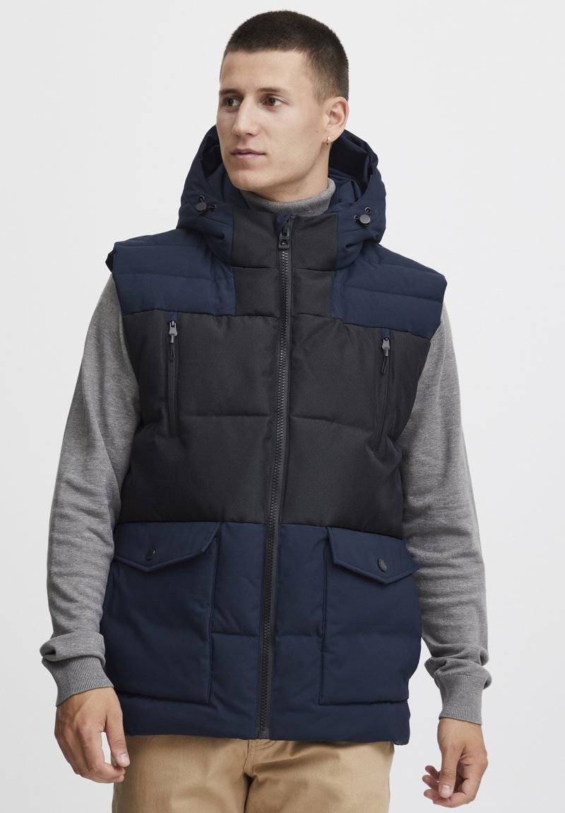 Navy and black puffer vest with a hood, zippered pockets, and buttoned front, worn over a gray long-sleeve shirt. Textured fabric.