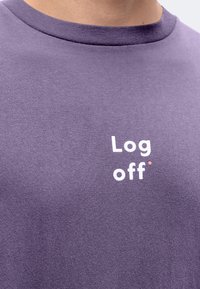 Човек, носещ лилава риза с бял надпис "Log off" на гърдите. Ризата има кръгло деколте.