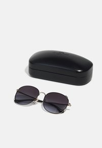 Óculos de sol com lentes redondas e escuras e armações de metal dourado, colocados ao lado de uma caixa rígida preta com superfície lisa e logotipo em cima.