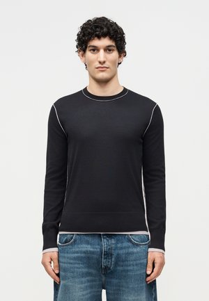 Giovane uomo con capelli ricci che indossa un maglione nero a maniche lunghe con dettagli di cuciture bianche e jeans blu, in piedi di fronte a uno sfondo bianco.