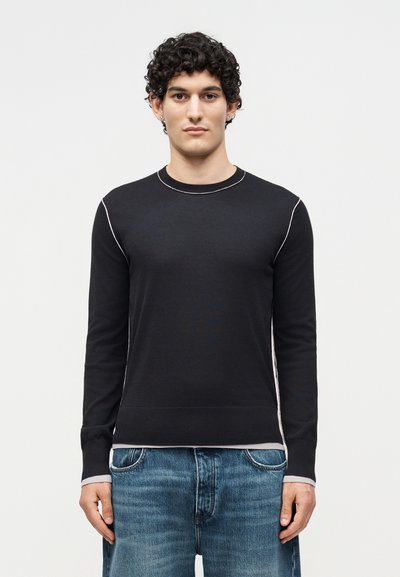 Jeune homme aux cheveux bouclés portant un pull noir à manches longues avec des accents de coutures blanches et un jean bleu, debout devant un fond blanc.