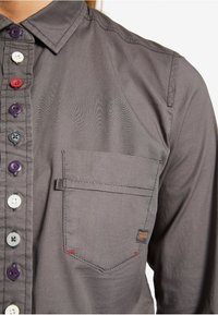 Grå skjorte med knapper og krave, med flerfarvede knapper, venstre brystlomme og diskrete syningsdetaljer på stoffet.