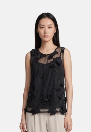 Donna che indossa un top nero trasparente senza maniche con applicazioni floreali sopra pantaloni beige su sfondo semplice.