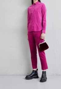 Chemise et pantalon en velours côtelé fuchsia à texture lisse, assortis à des bottines noires à semelle épaisse et un petit sac à main en velours rouge.