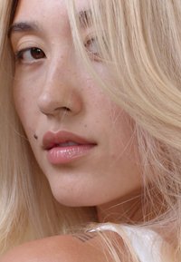 Gros plan sur un visage avec de longs cheveux blonds raides. Les traits incluent des yeux brun clair, des lèvres pulpeuses et une beauté marquée sur le menton.