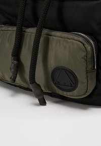 Sac à dos noir avec une poche avant rembourrée de couleur olive verte à fermeture éclair, un cordon de serrage noir texturé et un écusson circulaire noir.