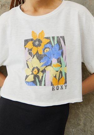 Frau trägt weißes, kurz geschnittenes T-Shirt mit buntem gelb-blauem Blumenmuster und "ROXY" Schriftzug, kombiniert mit schwarzem Rock.