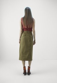 Kobieta z długimi prostymi włosami, ubrana w bezrękawnik w kolorze rdzy, oliwkową spódnicę midi z tylnymi kieszeniami oraz brązowe botki za kostkę, stoi na tle w jednolitym kolorze.