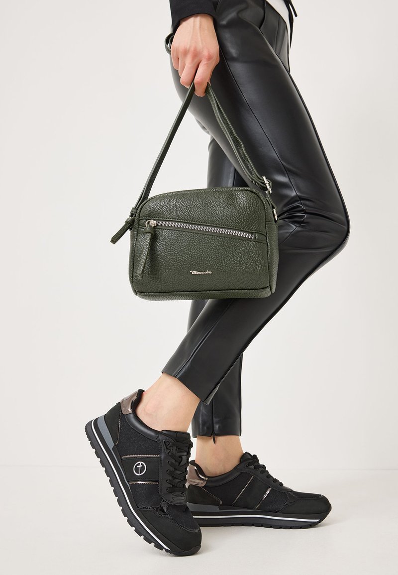 Green textured leather crossbody bag with two zippered pockets, held in a hand. Black fitted faux leather pants and black sneakers with accents.