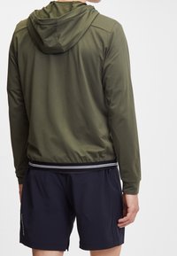 Giacca con cappuccio verde oliva, con vita aderente, texture opaca e accenti a righe bianche e nere all'orlo. Indossata sopra pantaloni corti blu navy.