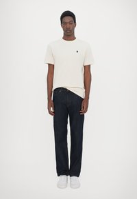 Beige bomull t-skjorte med en liten svart logo, kombinert med mørk blå denimjeans og hvite joggesko. Enkel, avslappet passform.