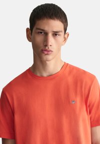 Oransje bomull t-skjorte med rund hals og korte ermer, med et lite brodert logo på venstre bryst. Glatt tekstur.