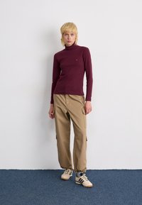 Maglione a maniche lunghe color marsala con collo a turtleneck abbinato a pantaloni cargo beige con tasche laterali e polsini elasticizzati. Le scarpe sono beige con dettagli neri.