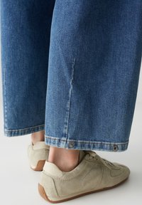 Grand pantalon en denim bleu avec jambes larges, détaillé par un léger effilochage à l'ourlet et des boutons-pression en métal. Associé à des baskets beiges.