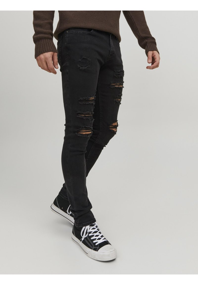 Svarta slitna skinny jeans med flera revor, en figurnära design och en mjuk textur, tillsammans med svarta och vita sneakers.