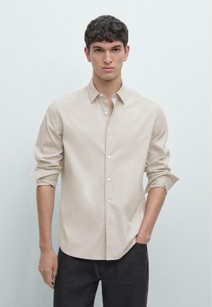 Giovane uomo con capelli ricci scuri che indossa una camicia beige con bottoni e le maniche arrotolate, e pantaloni neri, in piedi davanti a uno sfondo chiaro uniforme.