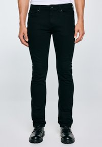 Mężczyzna w czarnych dopasowanych jeansach i czarnych skórzanych półbutach na sznurowadła, stojący na tle jasnego, jednolitego tła.