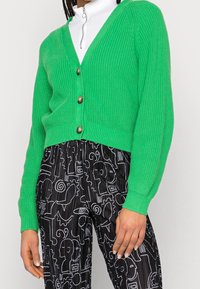 Person som bär en ljusgrön knappad kofta över en vit zip-up-top och svarta byxor med ett mönster av vita abstrakta linjeteckningar av ansikten.